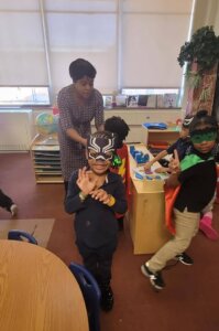 student dressed as a superhero poses for a picture in front of their teacher.